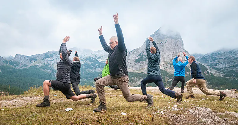Een groep mensen in outdoorkleding doet yoga of rekoefeningen op een grasveld, met op de achtergrond bergen en wolken.